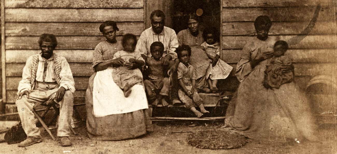 Black and white photo of an enslaved Africa-American family seated in front of cabin
