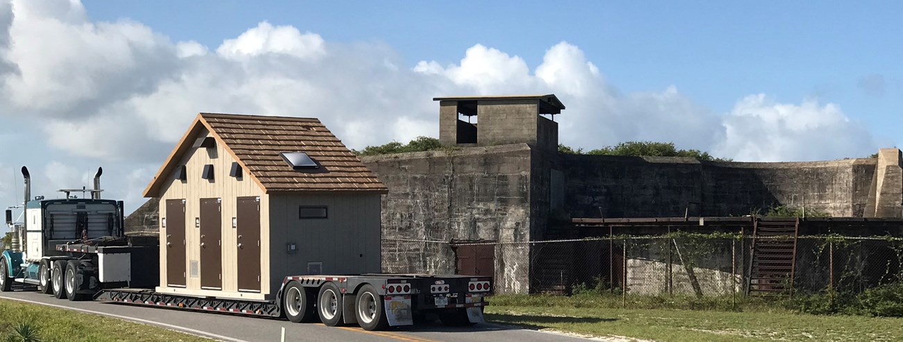 A semi-truck hauls part of a pre-fabricated restroom past a historic concrete gun battery. A semi-truck hauls part of a pre-fabricated restroom past a historic concrete gun battery.