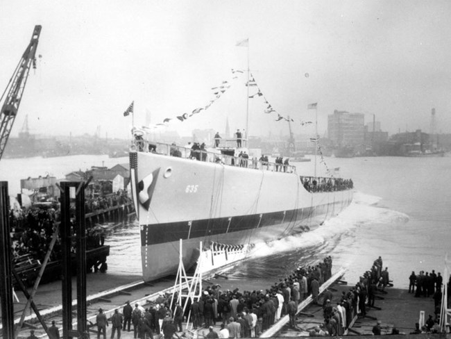 Photograph of a steel destroyer hull decorated in signal flags sliding into a harbor. Crowds on the shore and piers watch. City in the background.