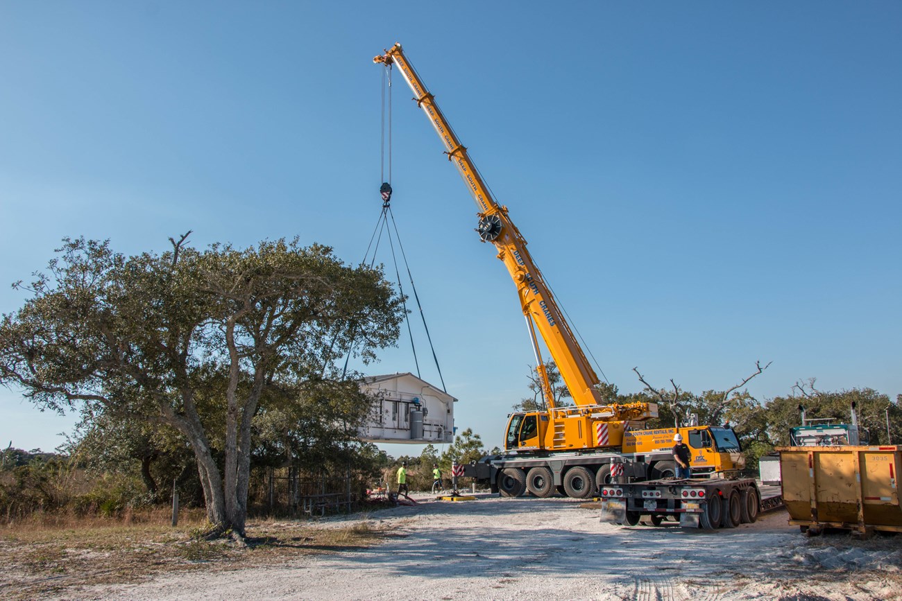A crane lifts a pre-constructed building into place in a sandy area.