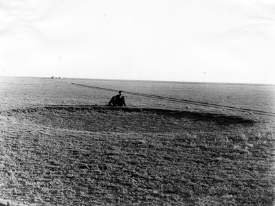 A black and white photo of a man sitting behind a giant hole in the prairie left by a bison wallow