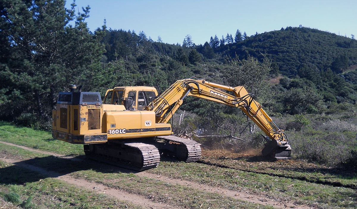 lt= An excavator digs off the side of a dirt road/trail