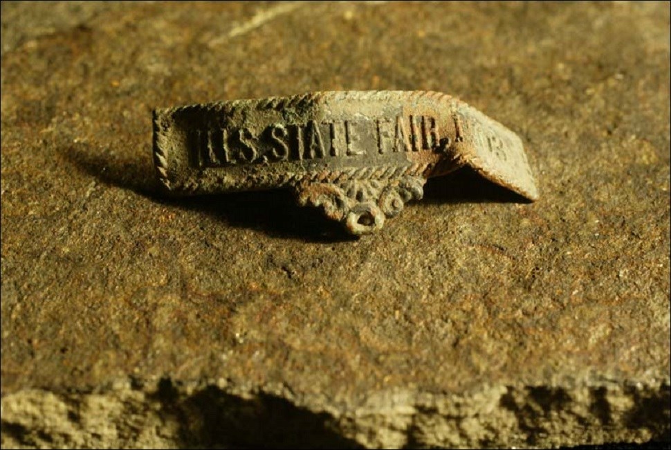 Piece of metal with words "US State Fair, 1903". An archaeological artifact. (Courtesy [Springfield, Illinois] State Journal Register, Photographer: Shannon Kirshner)