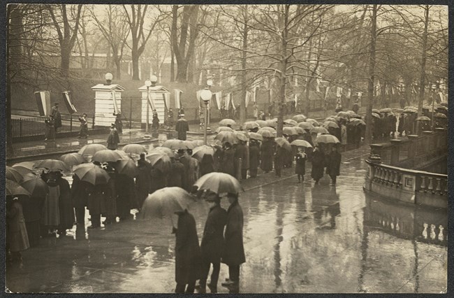 A crowd of people with umbrellas standing lining a city street; women holding banners visible in the background.