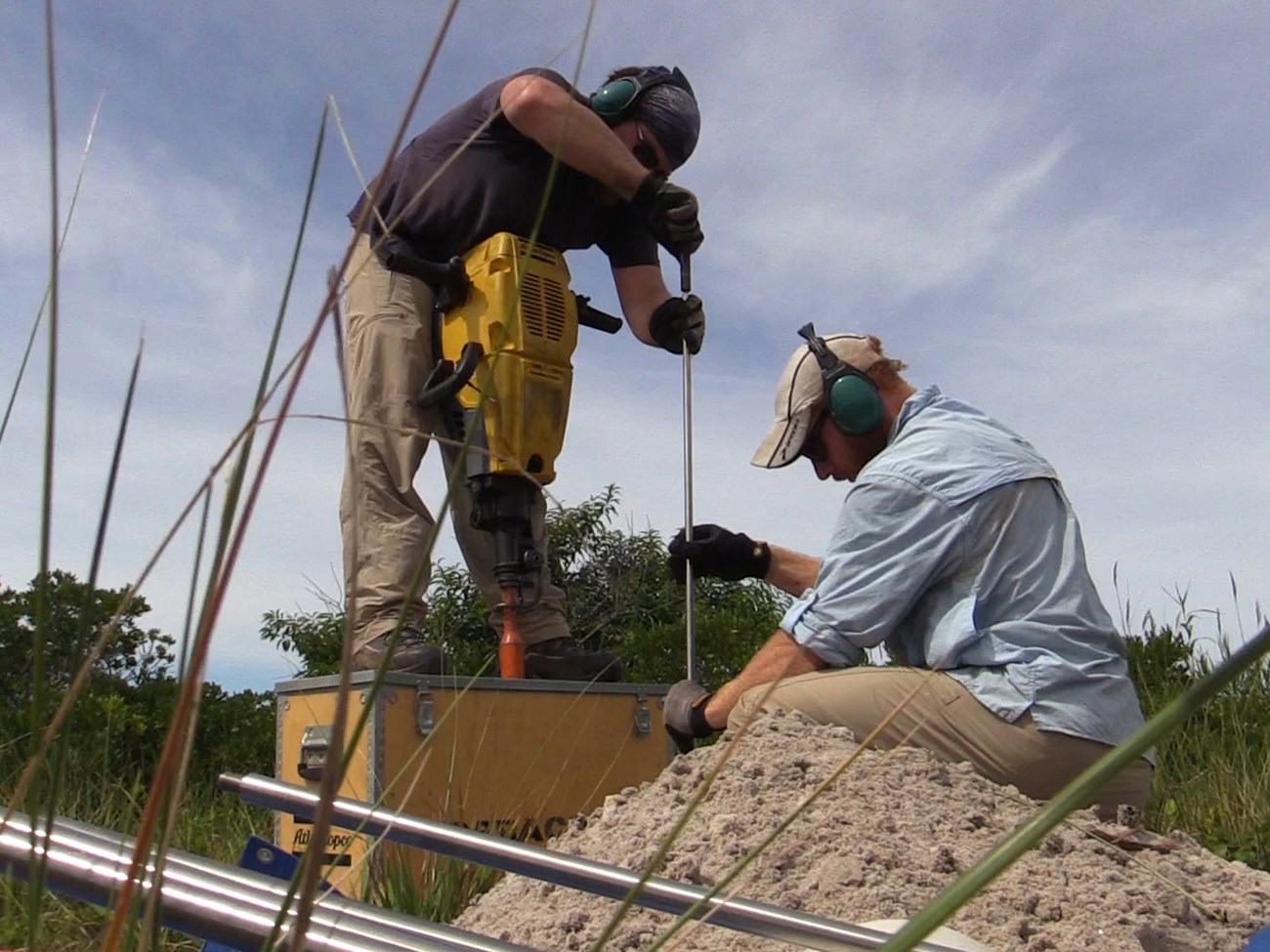 Two men use tools to install elevation markers in sandy soil.