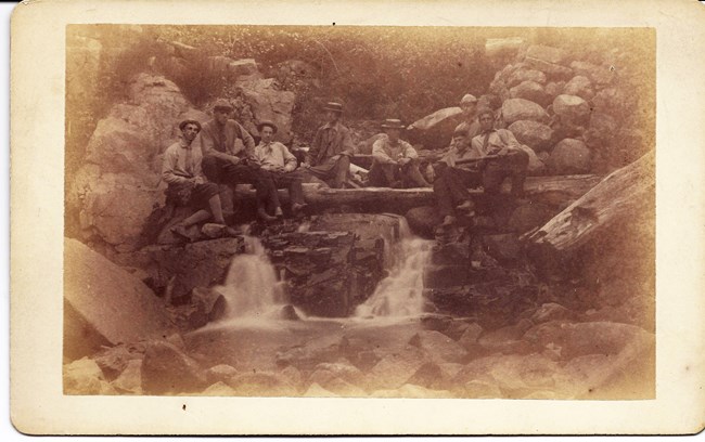 Eight men in a sepia-toned photo from the 1880s seated around a small waterfall.