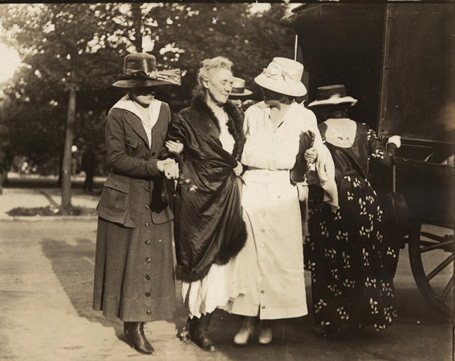 Woman wearing a coat over a prison uniform, walking assisted by other women.
