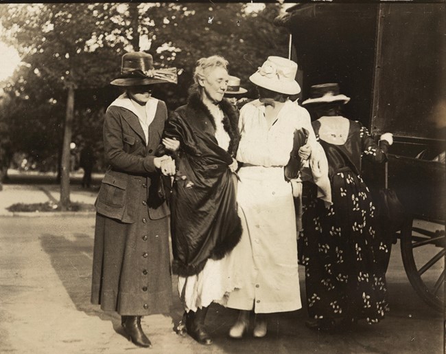 Dora Lewis on her release from prison. Woman wearing a coat over a prison uniform, walking assisted by other women.