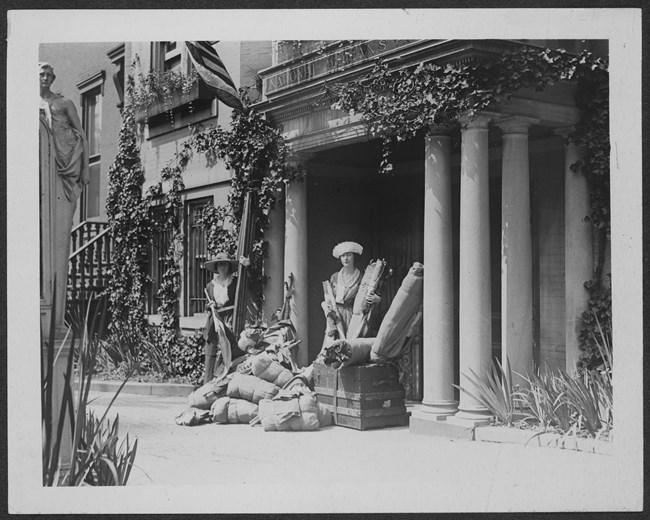 Two women stand with several suitcases and bundles outside a doorway with columns.