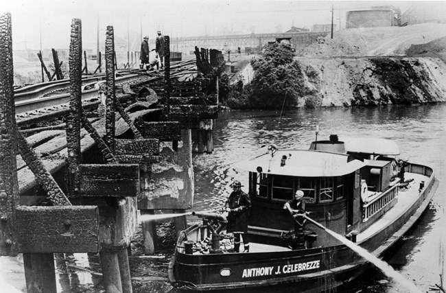 1969 Cuyahoga fire A black-and-white photo shows two firefighters spraying water from their boat up onto a burnt railroad bridge.