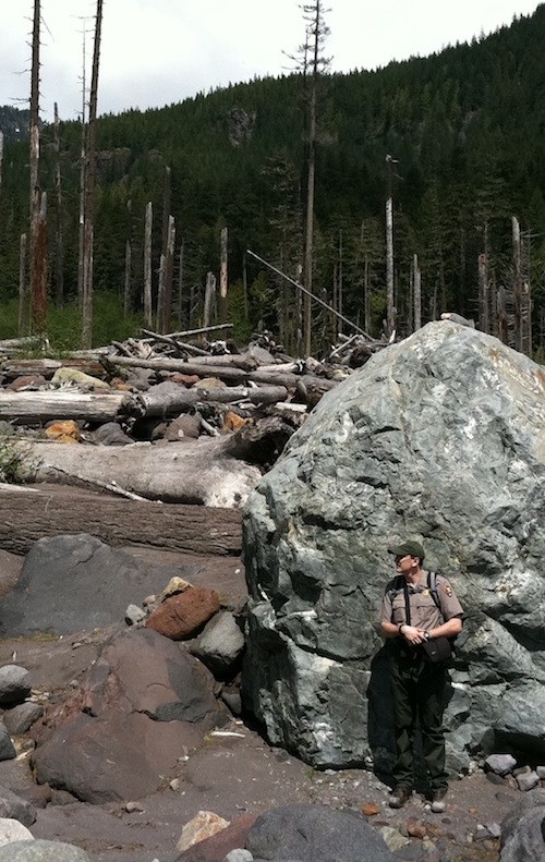 Boulders, logs, and other debris along Tahoma Creek. A ranger leans against a large boulder surrounded by tumbled logs and broken tree trunks.