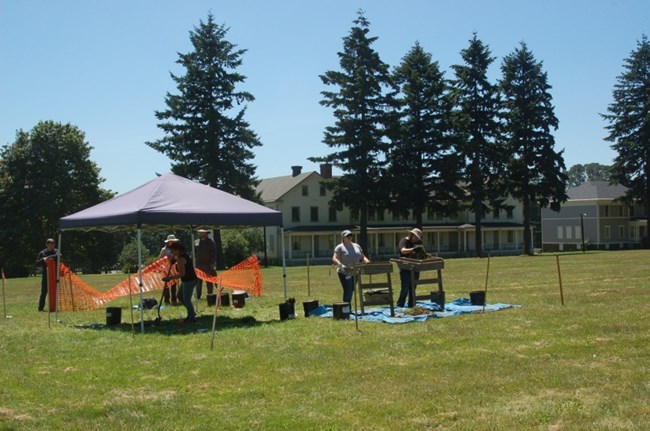 Two students screen dirt for artifacts while others work under a tent.