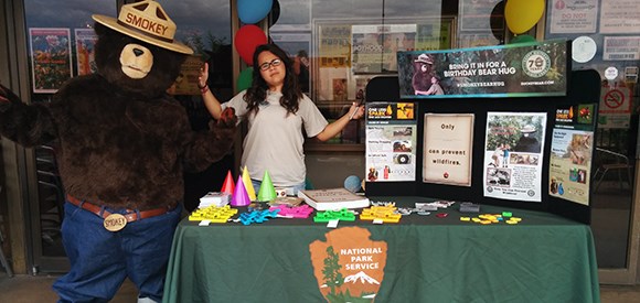A ranger and Smokey Bear stand behind a table with fire prevention materials.