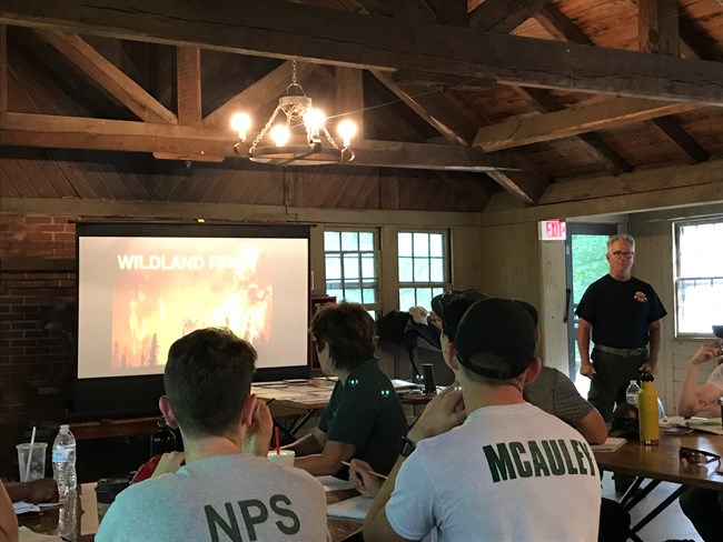 Instructor lectures in classroom in front of screen with headline about wildalnd fire.