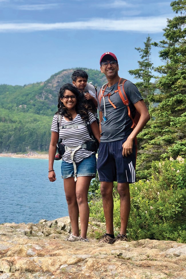 Family stands on rocky shoreline with beach and mountain in background Family stands on rocky shoreline with beach and mountain in background