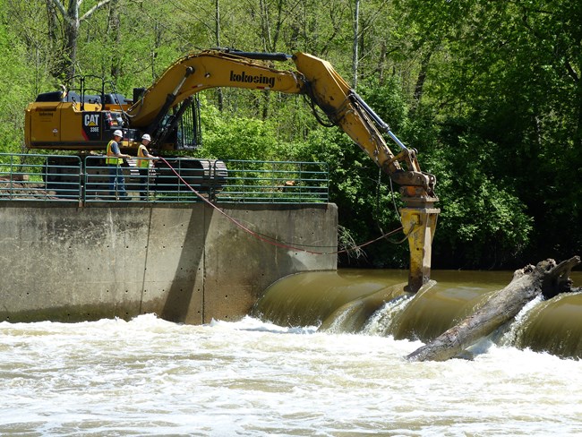 Two workers in vests and hard hats stand on shore as they operate a large yellow machine with its tip in the water over the dam.