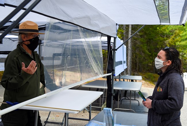 A park ranger and visitor have a conversation using social distance guidelines, with masks and a plastic barrier under an outdoor shade tent. A park ranger and visitor have a conversation using social distance guidelines, with masks and a plastic barrier under an outdoor shade tent.