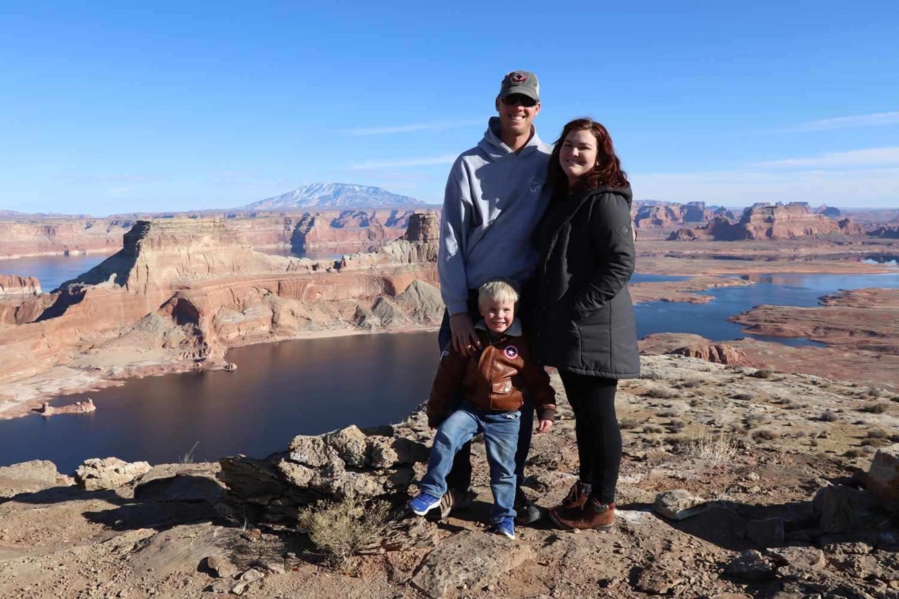 lt= Two parents and a child take a family photo near a scenic lake and canyon vista.