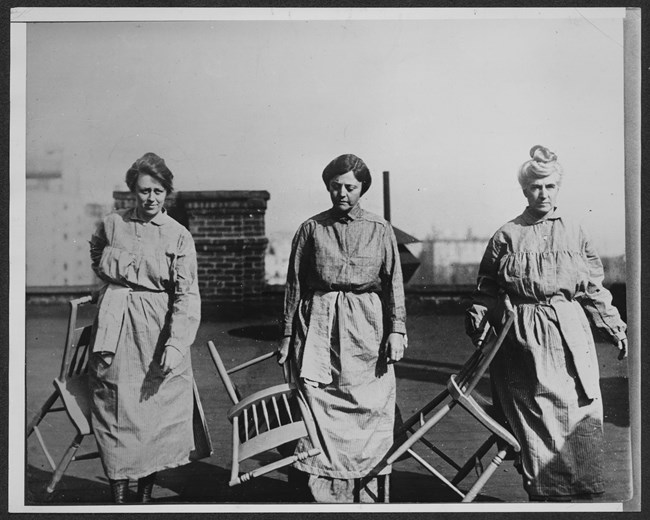 Three women dressed in prison uniforms hold wooden chairs and walk toward the camera.