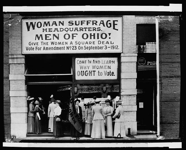 Several women stand outside a brick building with a sign heading "Woman Suffrage Headquarters. Men of Ohio! Come in and Learn Why Women Ought to Vote."