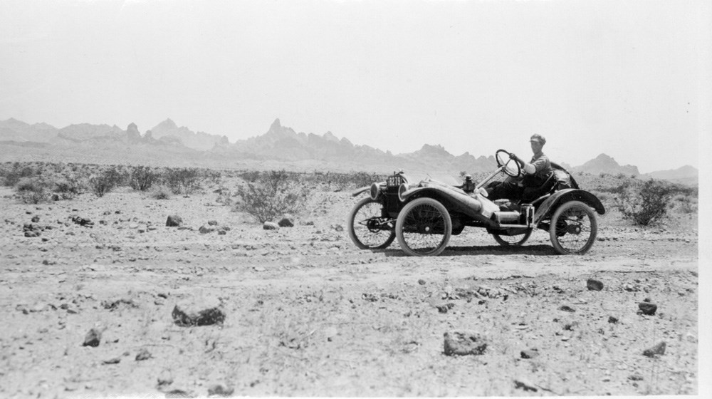 Driver operates car along a flat desert road. Rock formations can be seen in the distance.