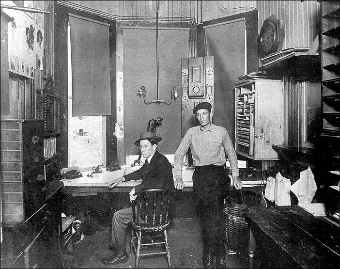 Two men at a desk, one sitting, one standing.