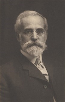 A bearded man with glasses in a suit looks at directly at the camera.