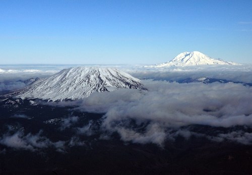 Mount St. Helens with Mount Rainier Two snowcapped volcanic peaks emerging above clouds.