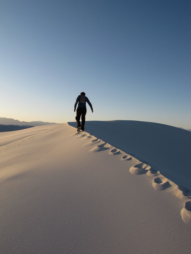 silhoetted person with a backpack hiking on the top of a dune ridge. Leaving tracks in the sand.