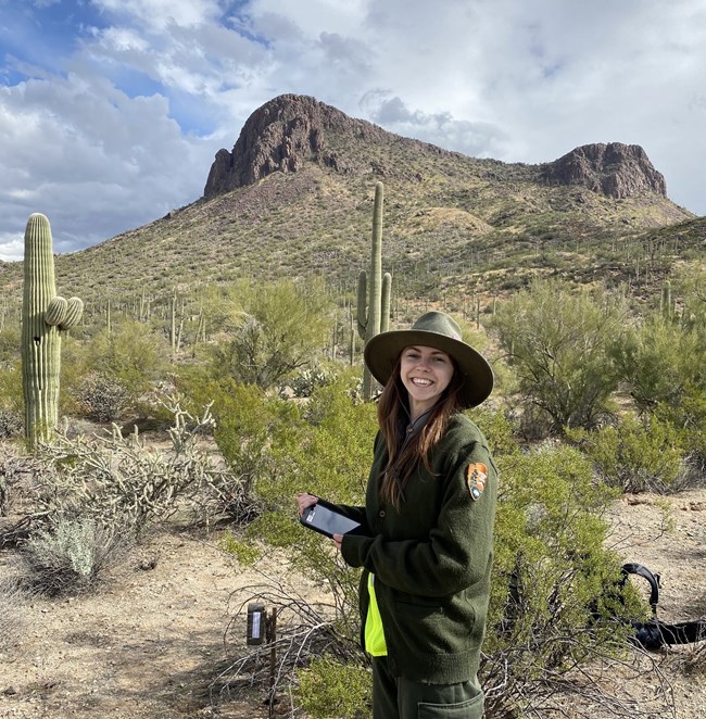 Person in NPS uniform stands next to a wildlife camera on a post.