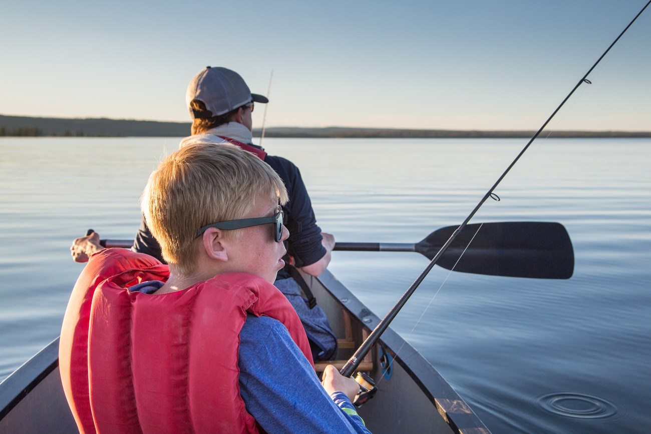 A young boy wearing a personal flotation device is holding a spin fishing rod in a canoe on a lake.