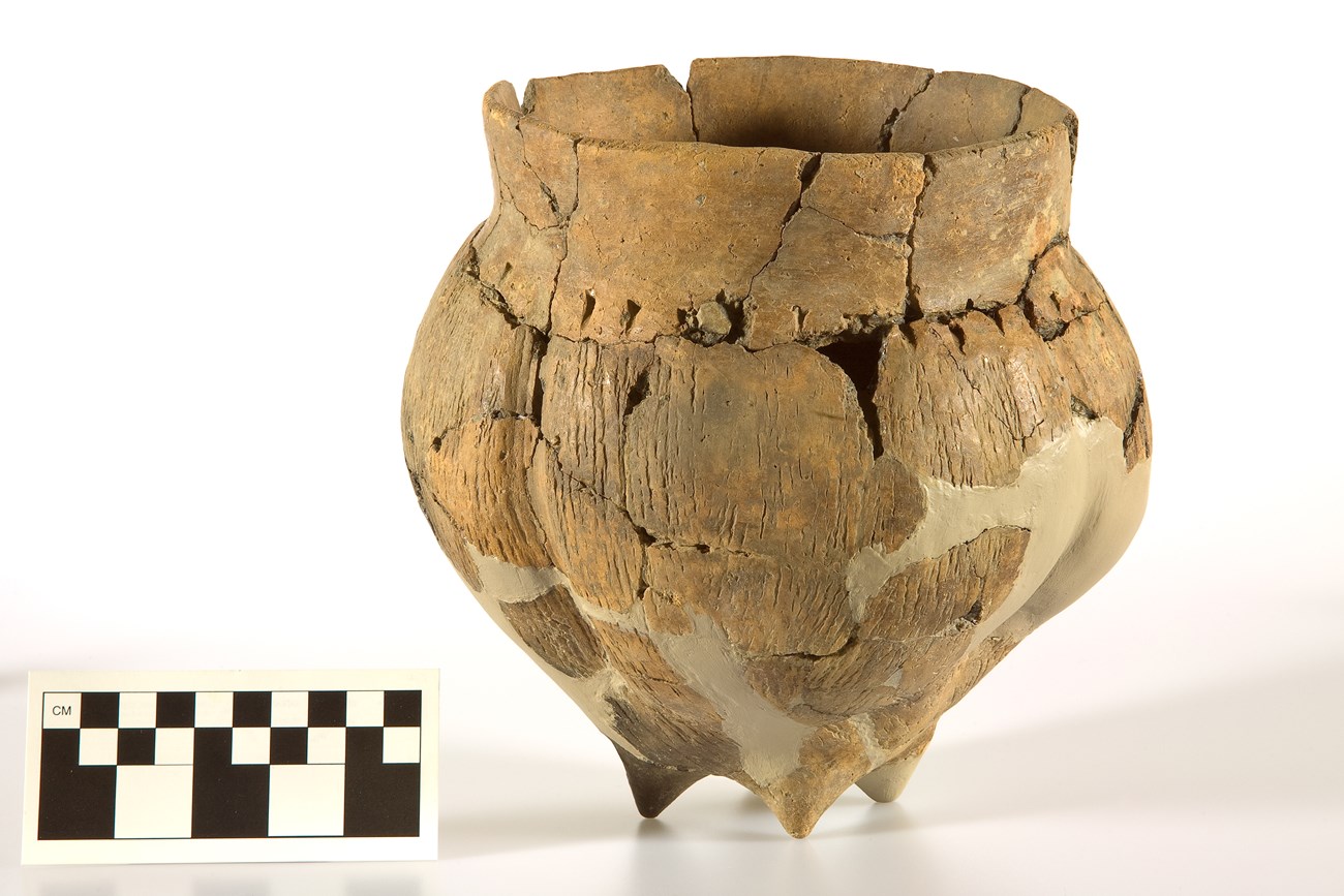 Clay pot with wide rim and vertical marks from cords. The pot has four small "feet" or pedestals.