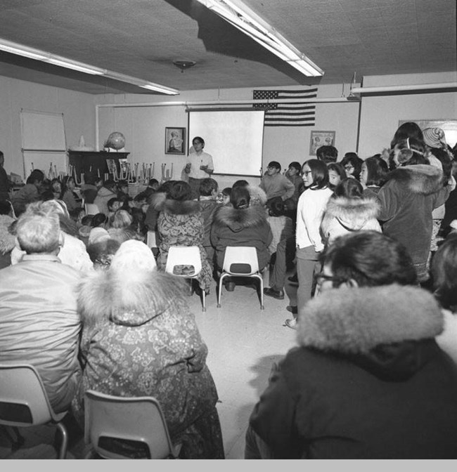 alaska native man speaking in front of a projector screen to a crowd of other alaska natives