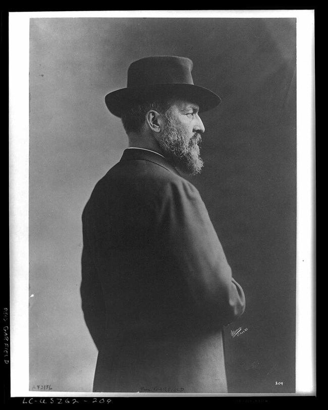 Black and white image of a James A. Garfield. He is facing away from the camera in profile. He wears a hat and long coat. He also has a full beard.