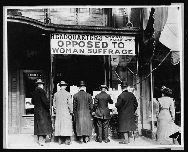 Men standing with their backs to camera under sign opposing women's suffrage. Library of Congress. Men standing with their backs to camera under sign opposing women's suffrage. Library of Congress.