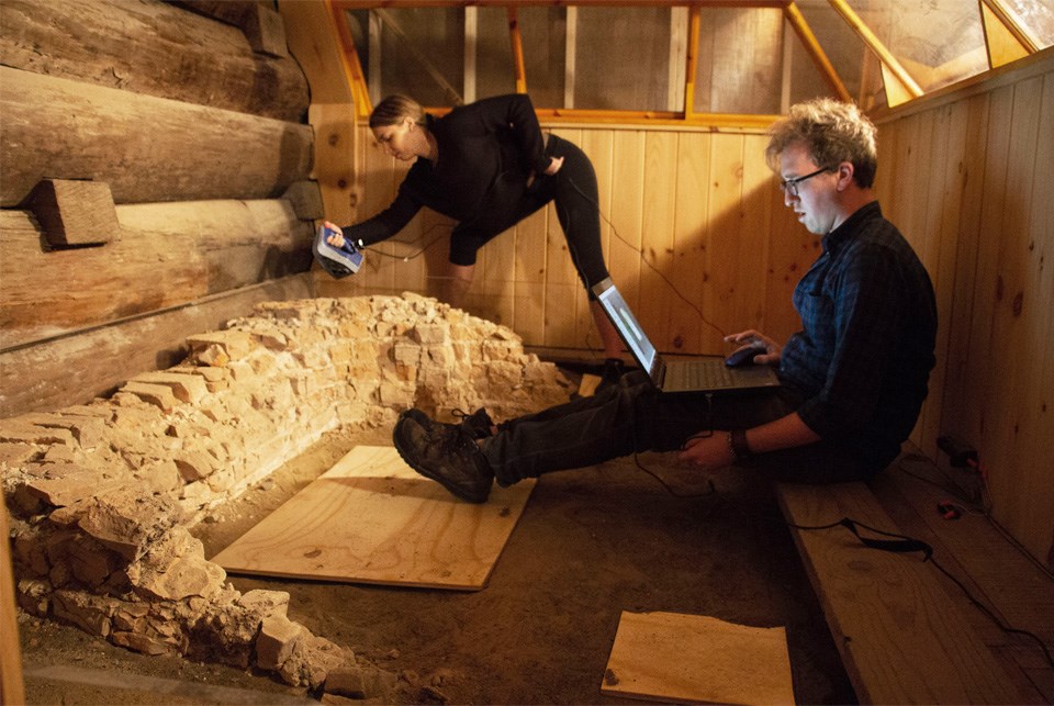 A man sits in a wooden boxy space with a laptop on his knees and feet near a crumbled brick hearth. A man sits in a wooden boxy space with a laptop on his knees and feet near a crumbled brick hearth.