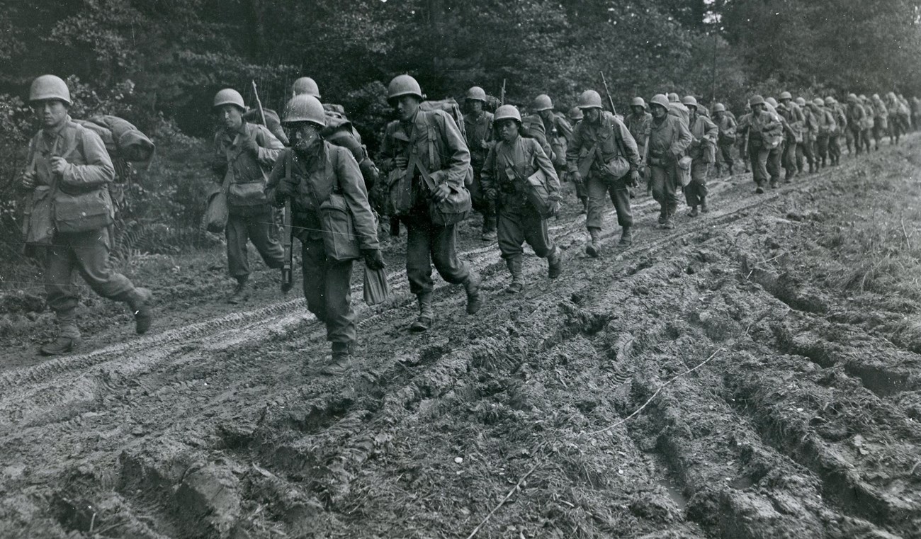442nd in France, 1944 A black and white photo shows a group of soldiers in uniform marching up a muddy path.