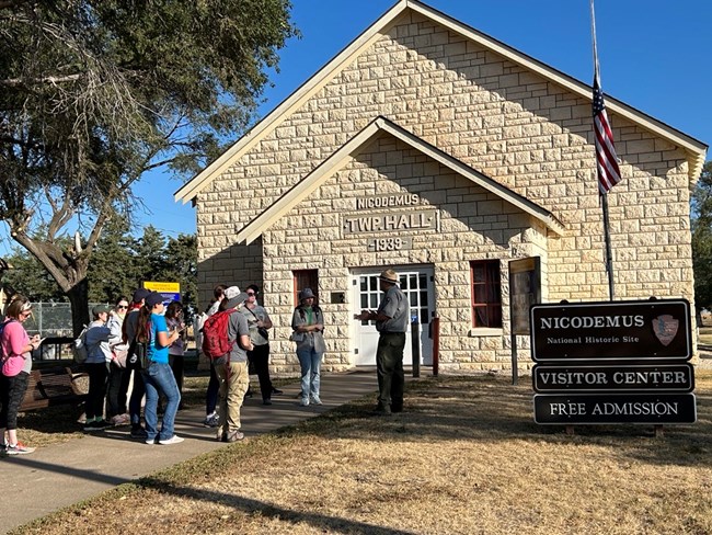 Visitors are always welcome to request town tours at Nicodemus NHS! NPS park ranger talking to a group of people in front of a an old limestone brick building. The building has white double doors with small window panes and a small window with red trim on either side of the door.