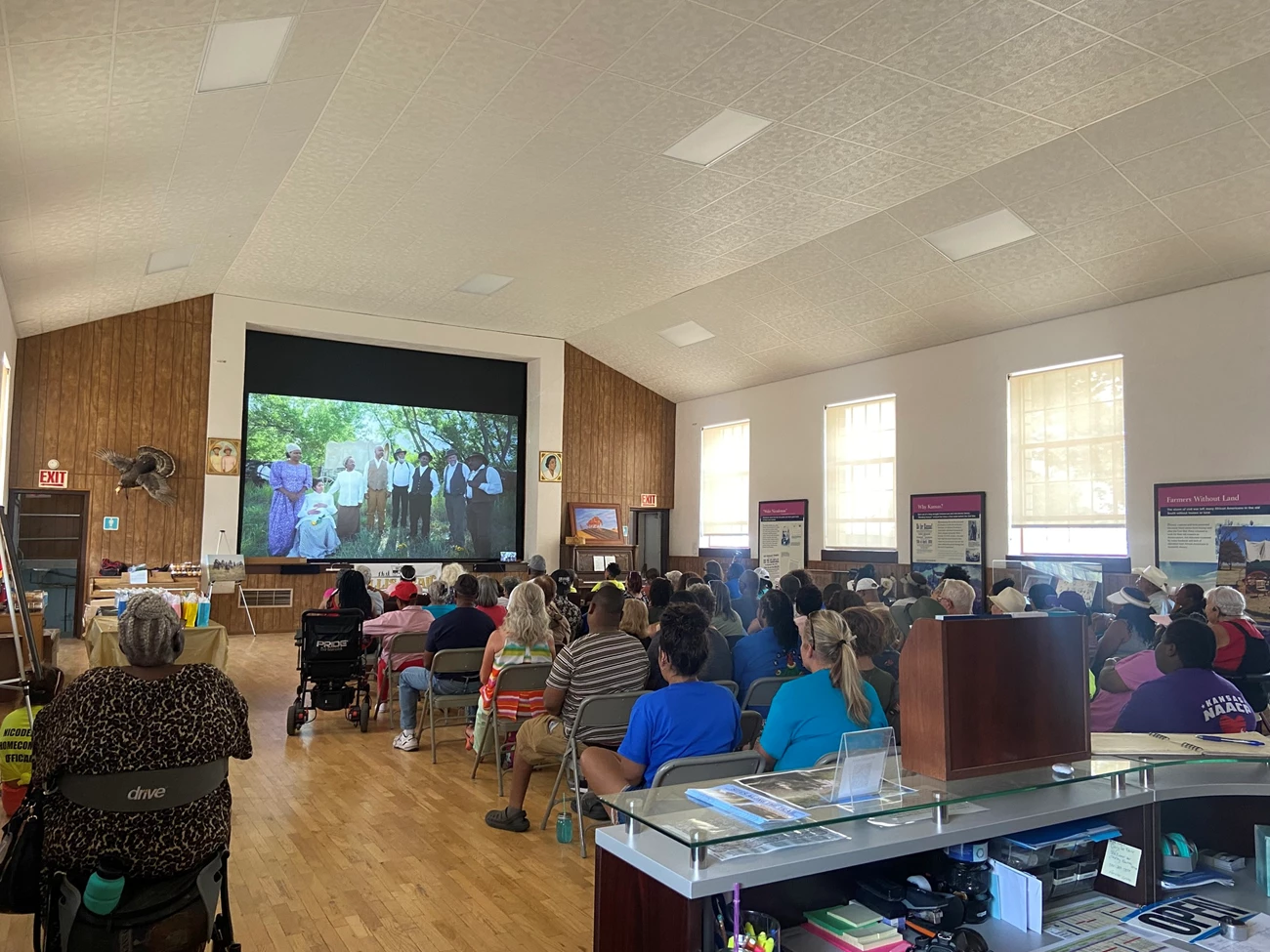 Nicodemus NHS has hosted a few showings of the 2024 documentary "Ellis Trail to Nicodemus." Photo shows the back of a crowd of people sitting in folding chairs and watching a film on a large projector screen. the image on the projector screen is a group of eight people dressed in clothing from the 1870s and standing in a forest clearing.