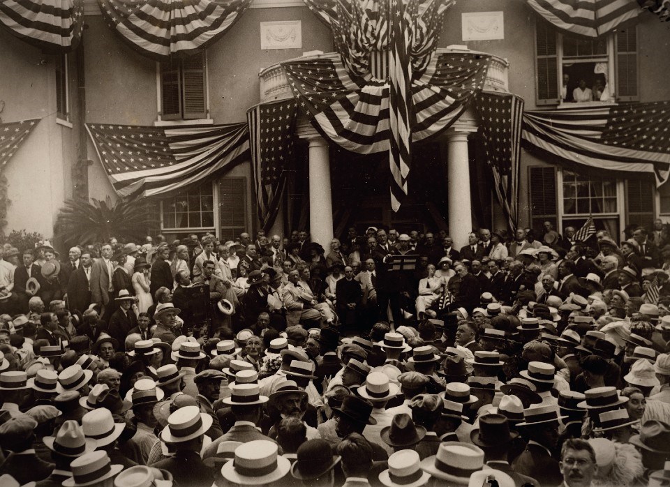 A crowd of onlookers attend a speech delivered from the portico of a house draped in United States flags.
