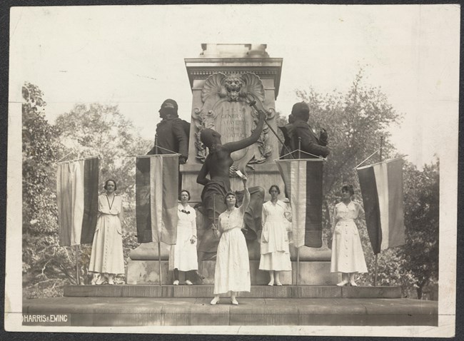Five women wearing white hold suffrage banners in front of a large monument.