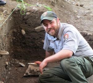 A man in a NPS uniform crouches in a dirt pit, smiling through a beard at the camera.