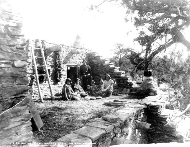 A traditional looking stone Hopi building with a small family of American Indians resting in the shade.