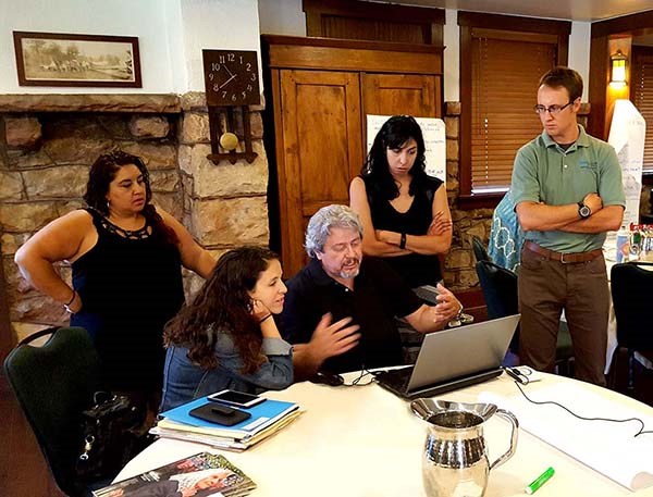 Five people stand and sit around a table looking at a laptop. Five people stand and sit around a table looking at a laptop.
