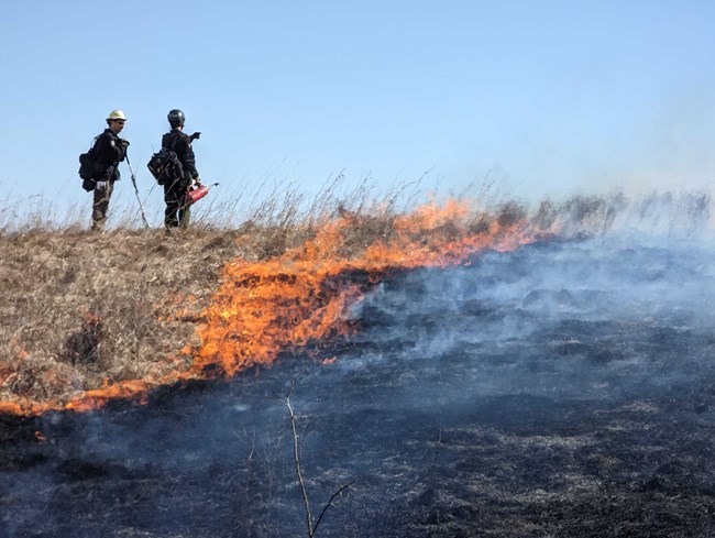 Two men stand in a prairie of dry brown grass feet away from an advancing wall of flames.