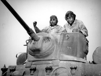Two men standing atop a tank behind the barrel of the mounted cannon