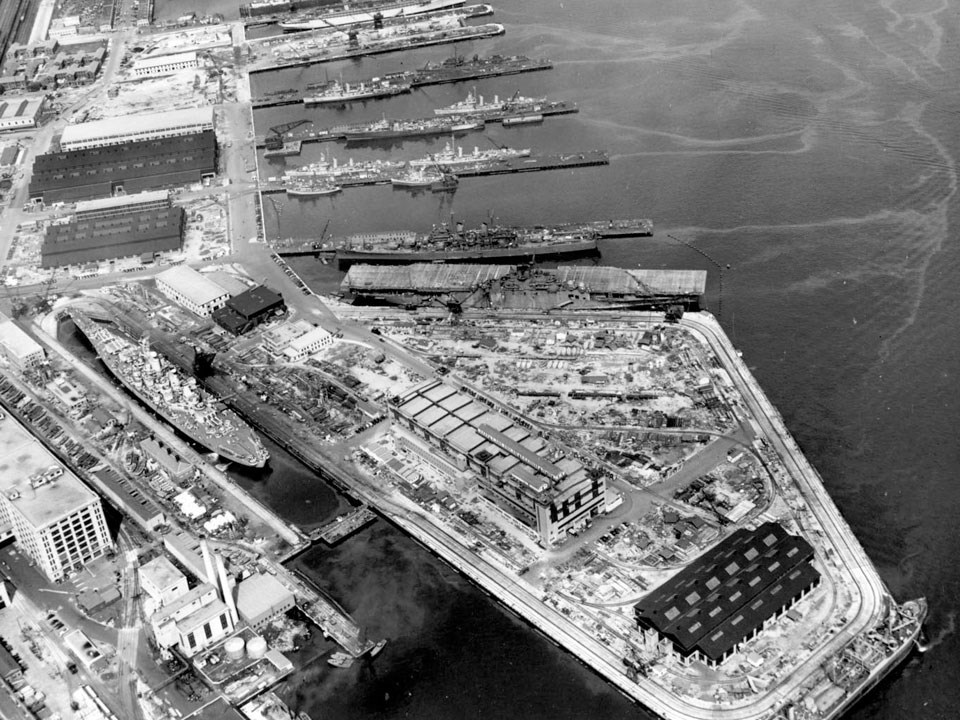 Photograph looking at piers dry docks and industrial buildings on the edge of a harbor. Warships are docked alongside the piers.