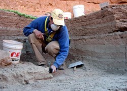 Archaeologist excavating one of the units. Archaeologist in a geometric hole about two feet down, excavating the earth.