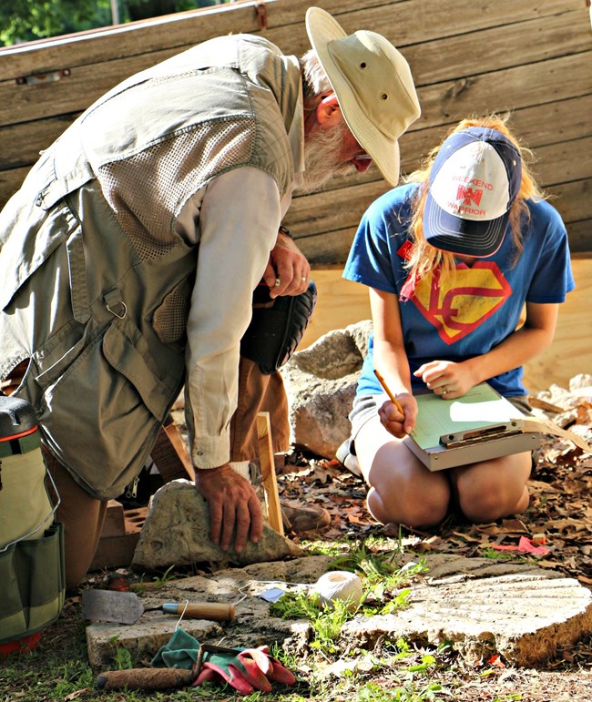 Man wearing a fishing hat kneels over a girl with a ball cap writing on paper.