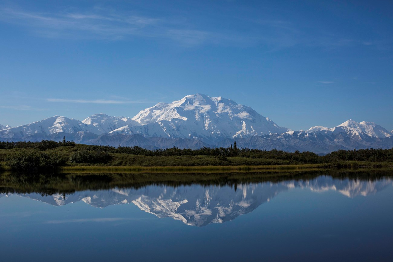 snowy mountains, one of which vastly dwarfs the others, in front of a lake and forested landscape